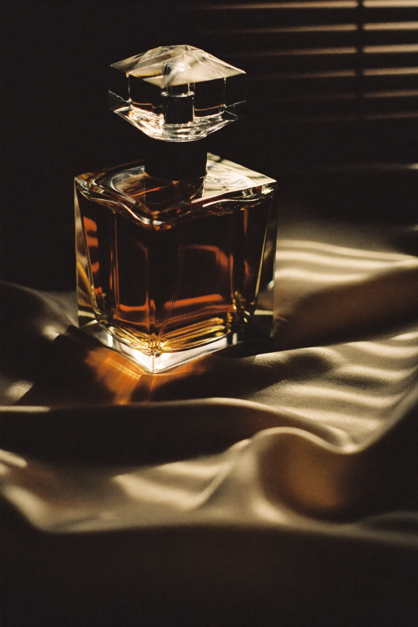 A faceted glass decanter of dark amber extrait standing on champagne silk, lit through venetian blinds.